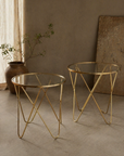 The Aria Brass Table, a pair of round glass-top side tables on looping gold brass legs beside a clay vase
