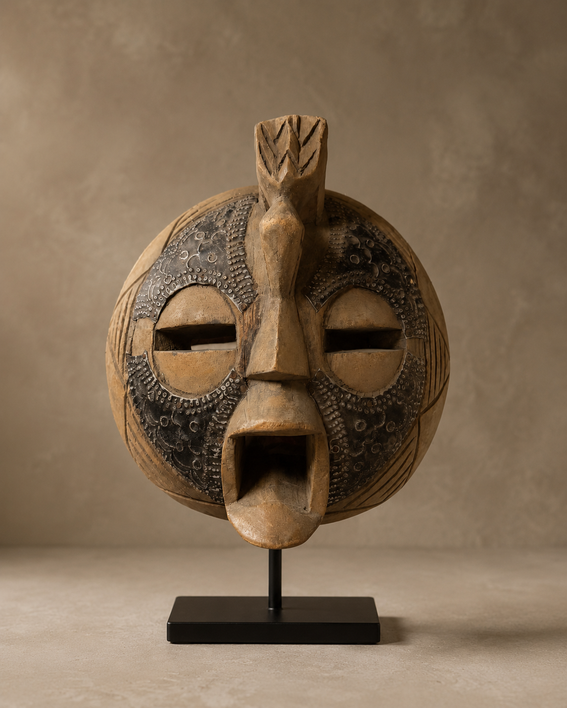 Ancient African Mask No.10, a round carved wood mask with hammered metal overlay and crest, on a black stand