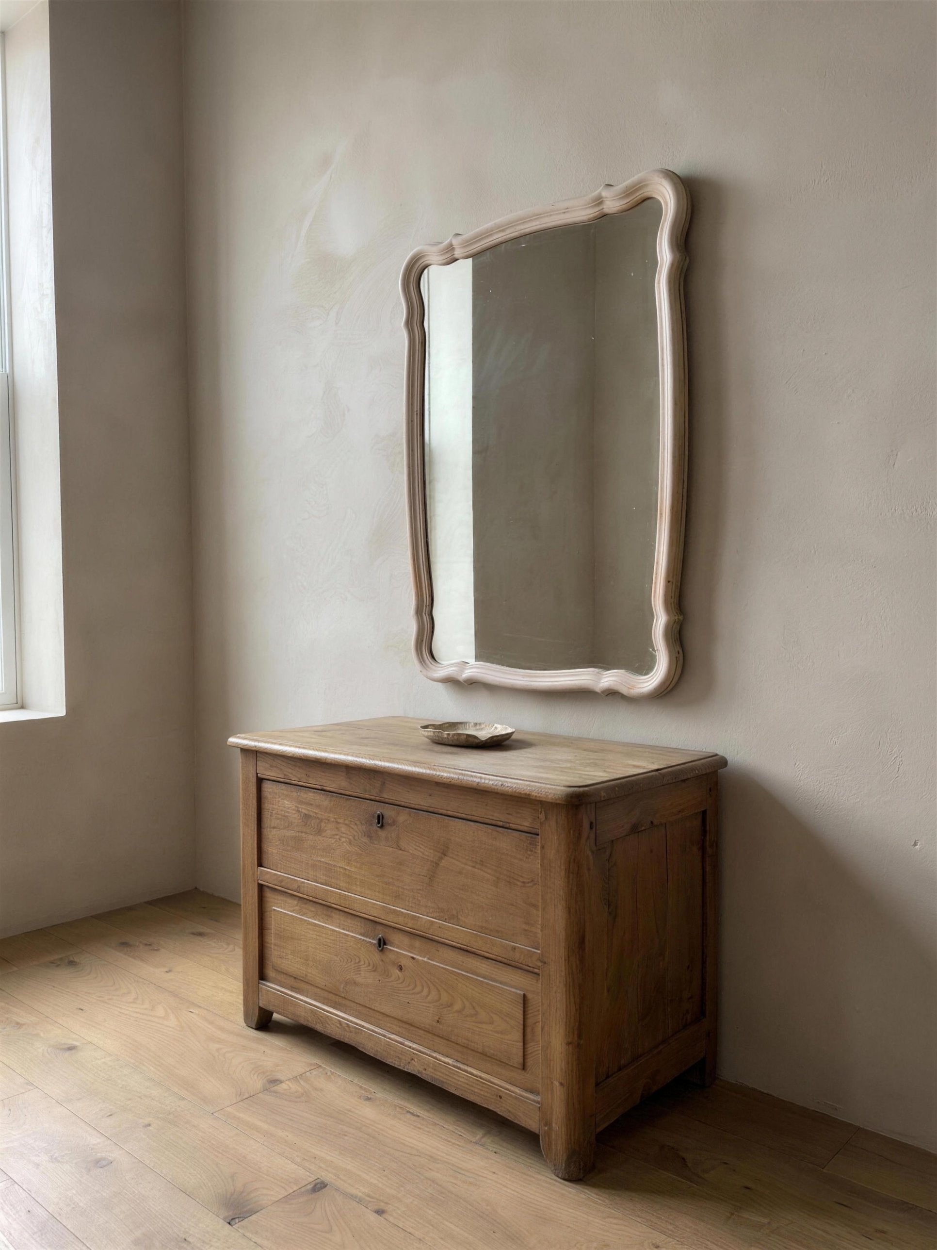Blush serpentine mirror hung above an antique wooden chest beside a window, seen at an angle