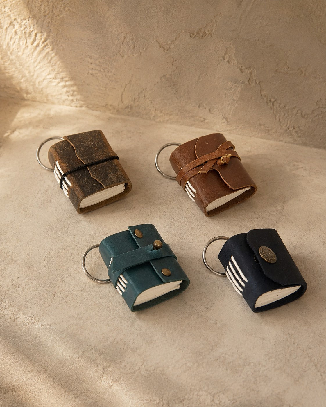 Four miniature leather journal keyrings in brown elastic, brown strap, teal and blue button, on travertine