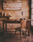 The Hawthorn Desk, antique wooden desk with a single drawer in a rustic plaster and brick room, primitive wood chair, faded rug, tall window light