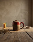 Lidded stoneware mug with red glaze breaking into warm sand, round loop handle, on a weathered oak table
