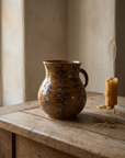 Speckled stoneware pitcher vase painted with columns of dark leaves, small red handle, on a weathered oak table