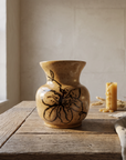 Speckled honey-toned vase with flared rim and a hand-painted dark bloom, on a weathered oak table