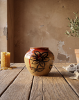 Round speckled stoneware vase with deep red glaze pooling at the shoulder and a painted dark flower, on a weathered oak table