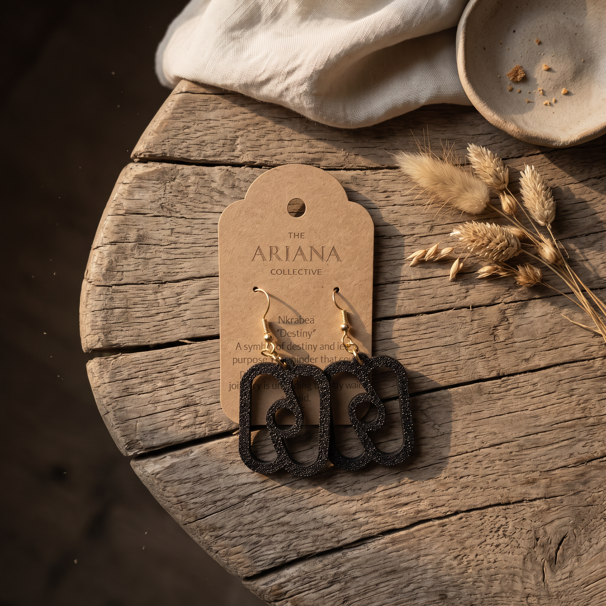 Nkrabea Adinkra earrings in black on a kraft Ariana Collective card, on a weathered oak table