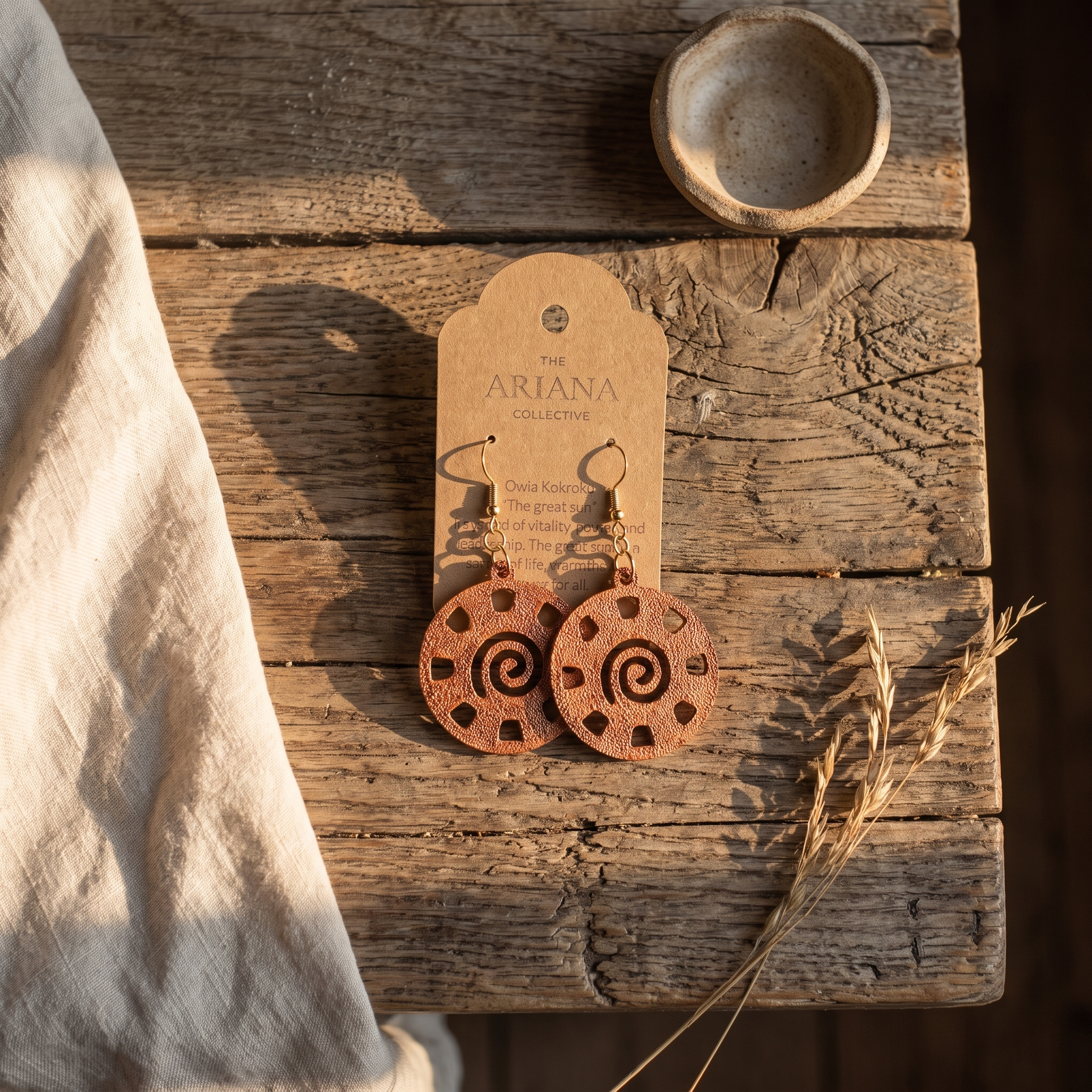 Owia Kokroko Adinkra earrings on a kraft Ariana Collective card, on a weathered oak table
