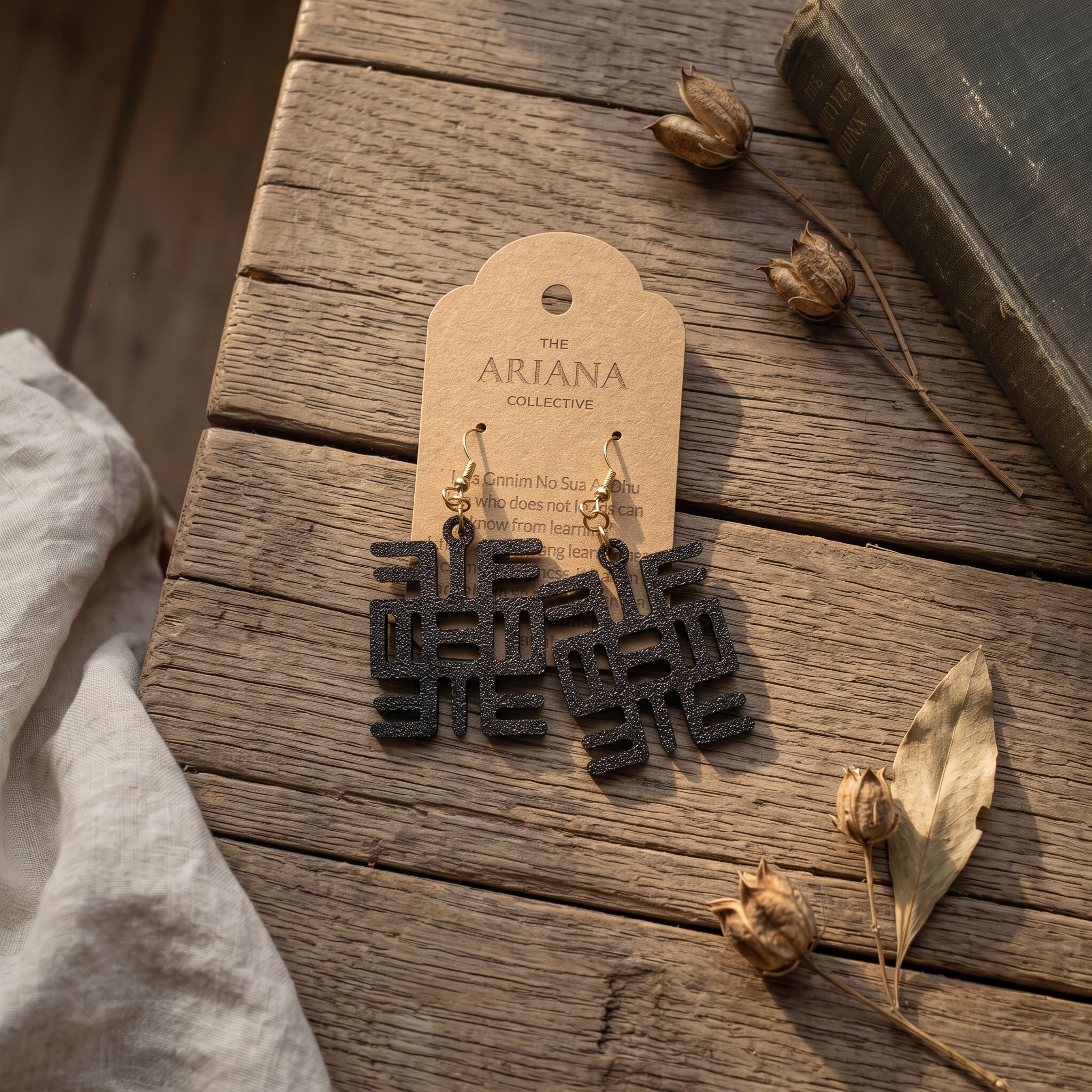 Nea Onnim No Sua A Ohu Adinkra earrings on a kraft Ariana Collective card, on a weathered oak table