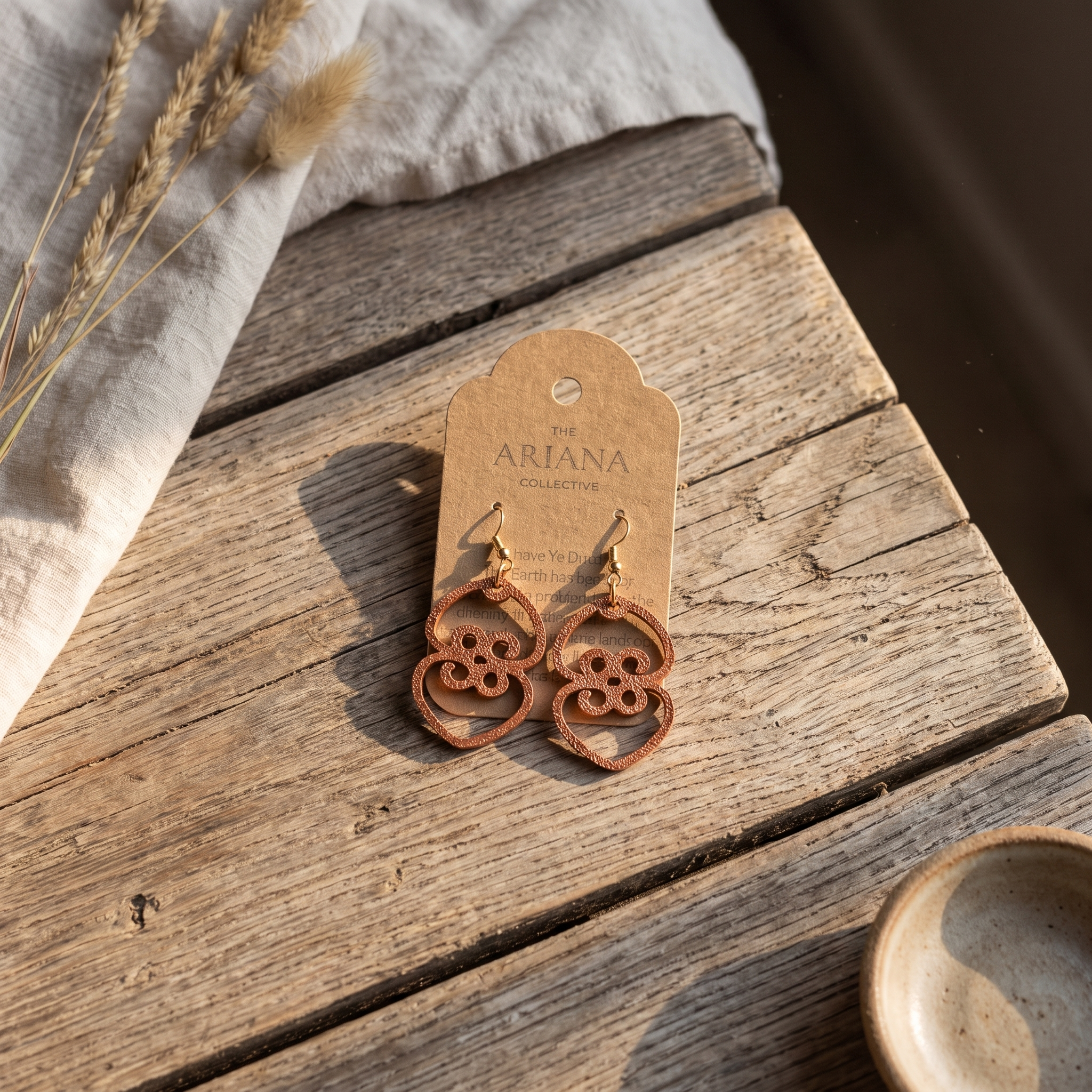 Asase Ye Duru Adinkra earrings in copper on a kraft Ariana Collective card, on a weathered oak table