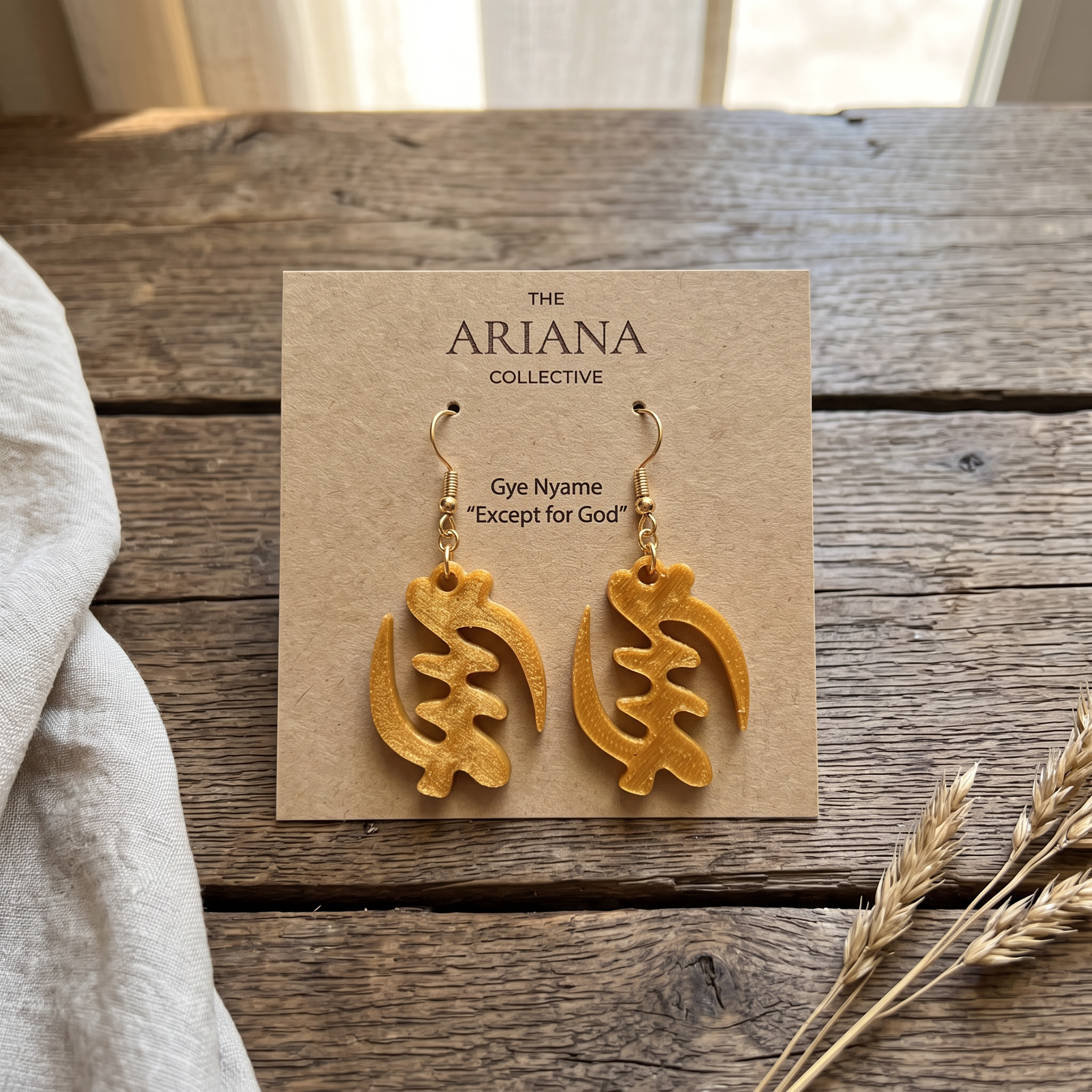 Gye Nyame Adinkra earrings in gold on their Ariana Collective card, laid flat on a weathered oak table