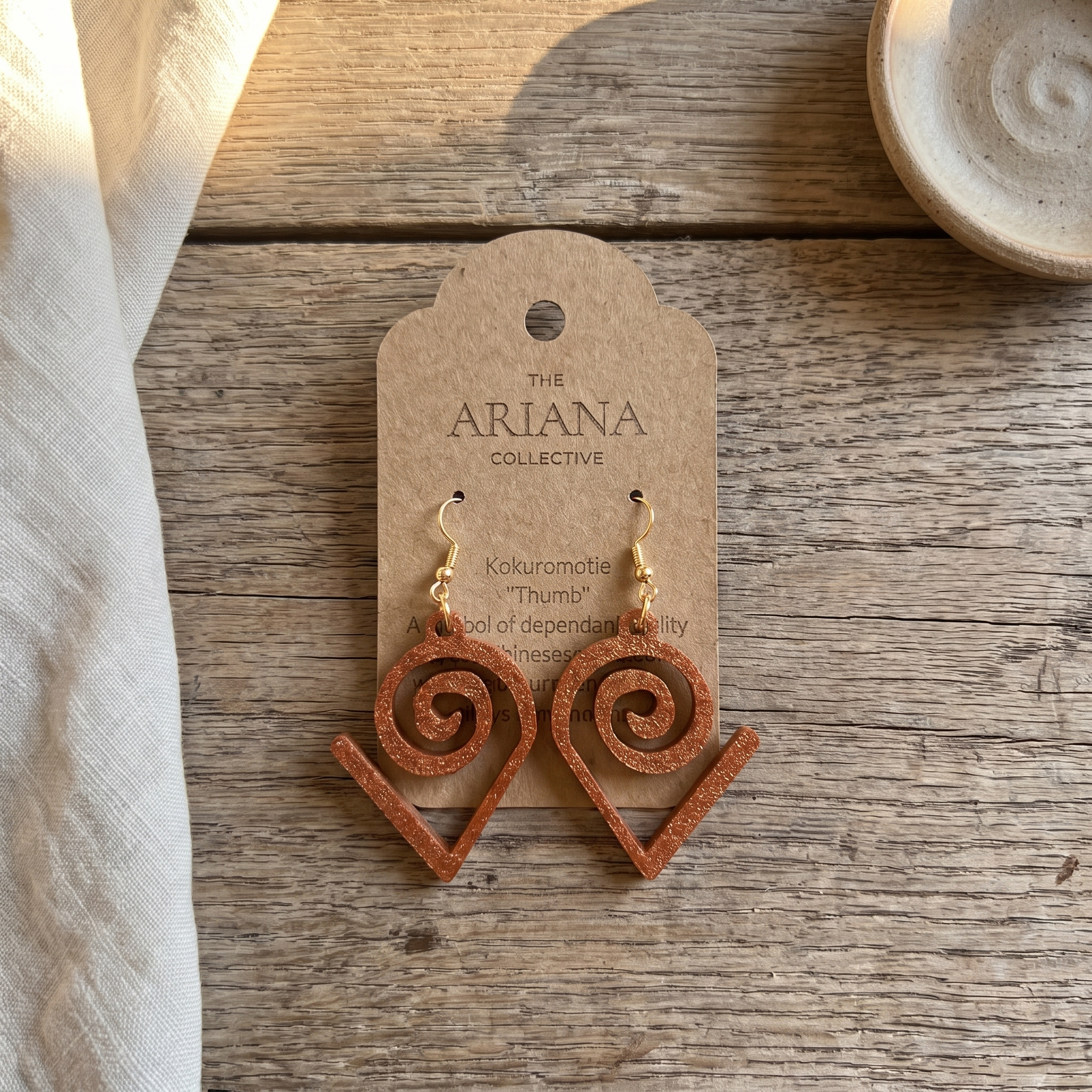 Kokuromotie Adinkra earrings in copper on their Ariana Collective card, laid flat on a weathered oak table