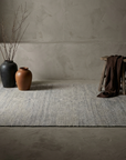 Heathered grey flatweave rug, lifestyle hero, on a greige plaster floor with stoneware vessels and draped linen
