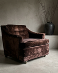 Three-quarter view of a single chocolate crushed velvet club chair showing the six-button tufted back cushion, rolled arms, upholstered plinth base and front caster