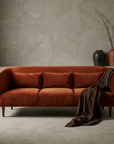 Burnt orange velvet curved sofa against a greige plaster wall, chocolate linen draped over the far arm, stoneware vessels and dried branches on a plaster ledge