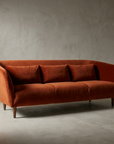 Three-quarter view of the burnt orange velvet curved sofa alone in an empty plaster room, showing the continuous curved shell and tapered walnut legs