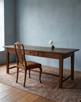 The Hawthorn Desk, a long low antique wooden writing desk with a carved walnut side chair upholstered in worn cognac leather pulled up to it, soft blue-grey plaster walls and a faded Persian rug on wide plank floors