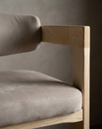Macro detail of the greige microsuede seat cushion, welted seam and the ash arm rail meeting the front leg at a sharp corner