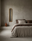 Slim arched giltwood mirror hung on greige plaster wall beside a low linen-dressed bed, terracotta vessel with bare branches on a plaster ledge
