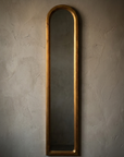 Straight-on view of the slim arched giltwood mirror alone on a bare greige plaster wall, worn gold leaf frame