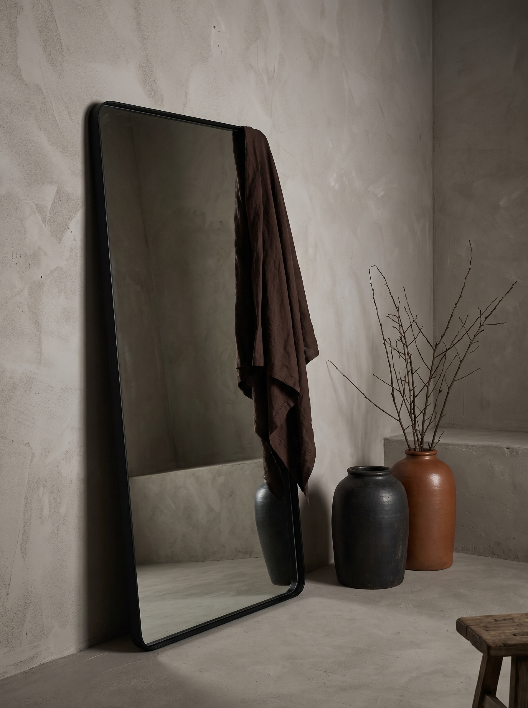 Large leaning floor mirror with matte black frame in a greige plaster room, styled with stoneware vessels and draped linen