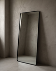 Three-quarter view of the large leaning floor mirror showing the rounded black metal frame