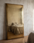 Brass panel mirror in a furnished bedroom corner, reflecting the bed and a side table