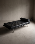 Barcelona Daybed three-quarter view showing full silhouette, tufted cushion, bolster, and legs against bare greige plaster
