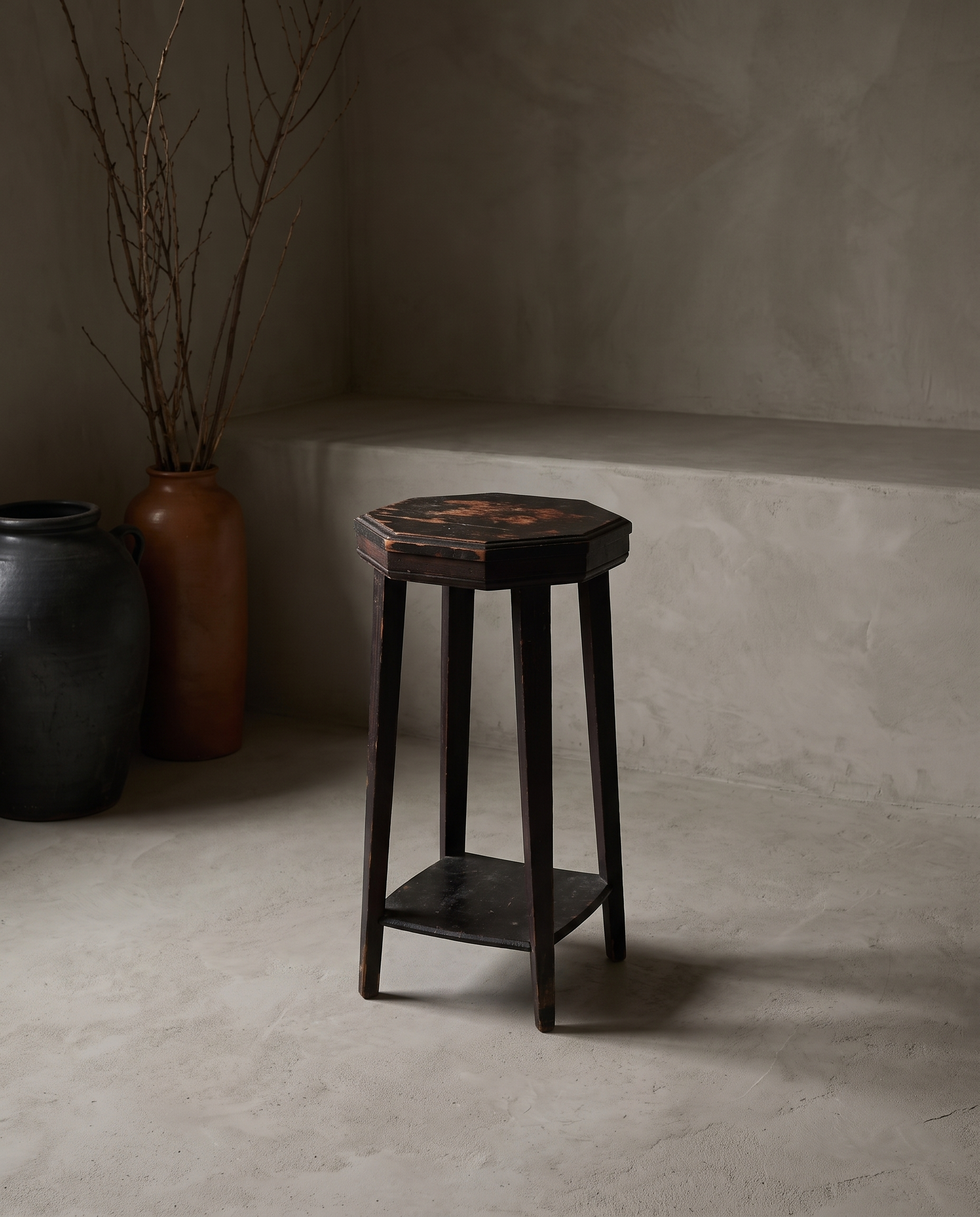Rustic accent stool with worn dark paint on octagonal top, styled with stoneware vessels and dried branches in a plaster room