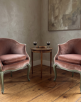 Pair of rose velvet bergères facing each other in a sitting room corner with a small wood side table, two glasses of wine and a book