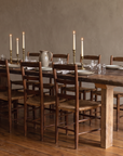Redwood dining table set for ten with rush-seat ladder-back chairs, brass candlesticks and a stoneware pitcher