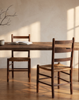 Redwood dining table in morning window light with a coffee cup, open book, branch in a stoneware vase and two rush-seat chairs
