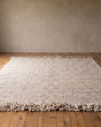 Awanay Jume Soga handwoven Criolla wool rug, 9 x 12, with shaggy wool fringe on both ends, laid flat in an empty room with plaster walls and a reclaimed wood floor