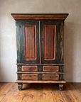 The Batavia Armoire, a painted green-black two-door armoire with red-brown panels and a four-drawer base, against a greige plaster wall on a reclaimed wood floor