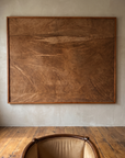 Untitled (Rust) by Nicole Daedone, a large framed work of rust and umber handmade paper hung on a warm plaster wall above a wood and suede armchair