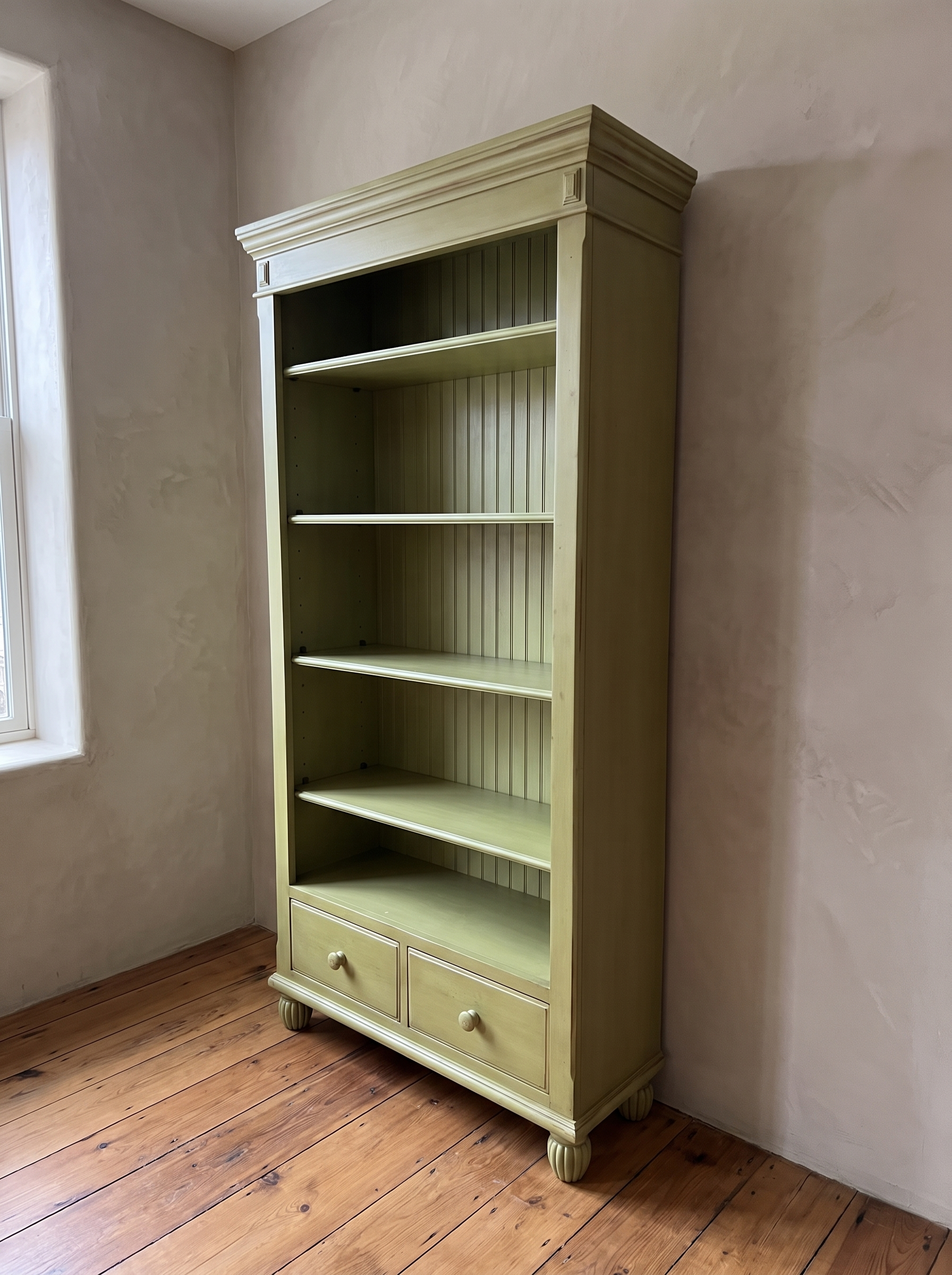 Three-quarter view of the olive green bookcase showing its plain side panel, drawers and reeded bun feet