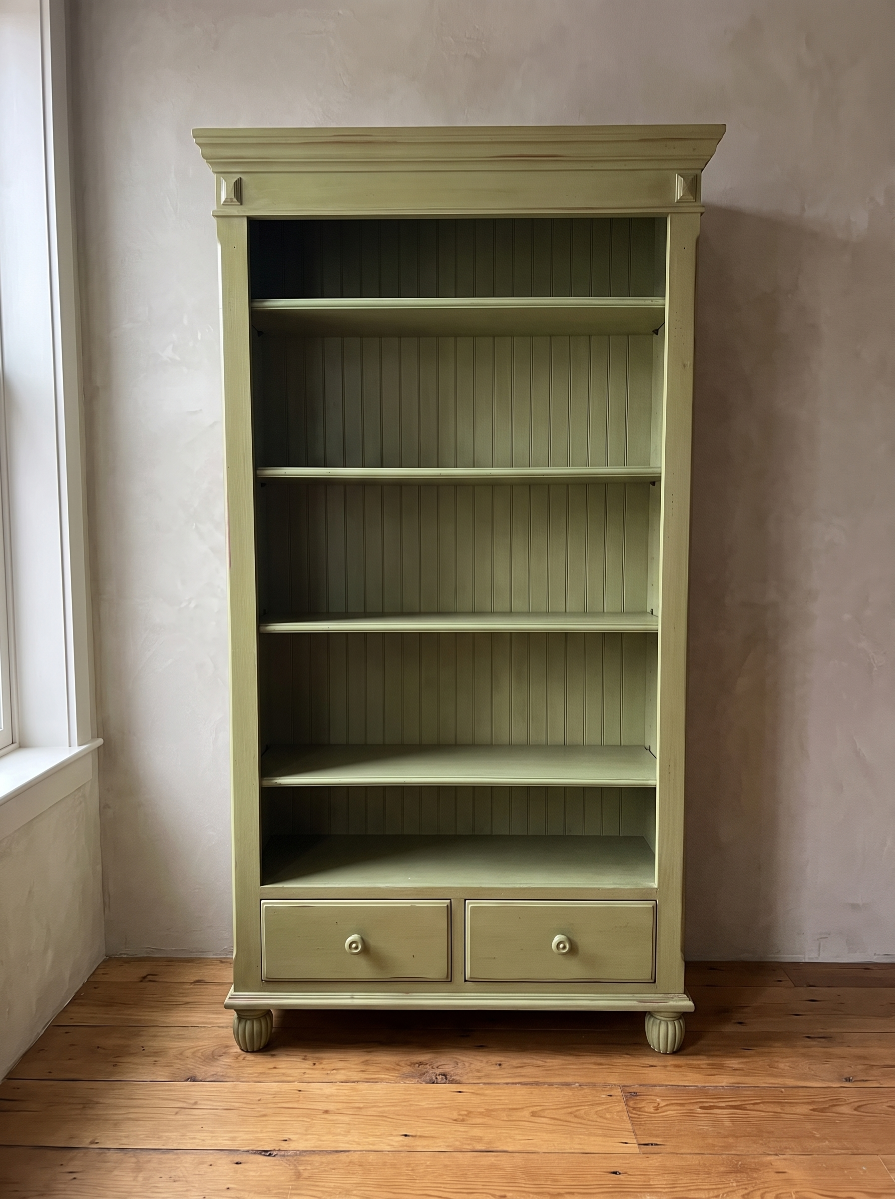 Olive green painted bookcase with cornice crown, beadboard back and two drawers, standing against a greige plaster wall on a wide-plank wood floor
