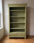 Olive green painted bookcase with cornice crown, beadboard back and two drawers, standing against a greige plaster wall on a wide-plank wood floor