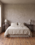 A matching pair of vintage Indian side tables on either side of a queen bed in oatmeal linen, a ceramic lamp on each, plaster wall and reclaimed wood floor