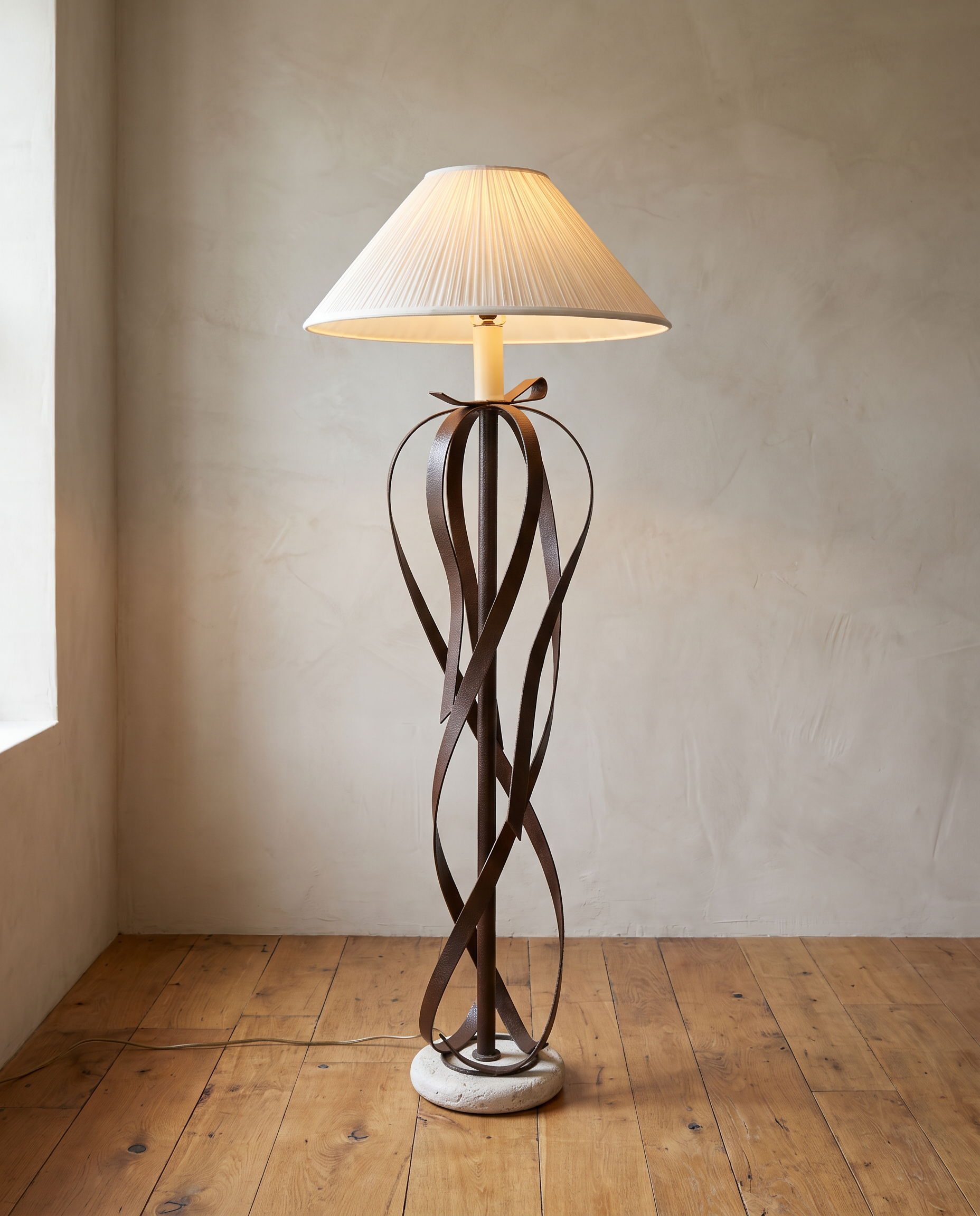 Ribbon floor lamp in hammered steel with a pleated shade, lit, against a plaster wall on a reclaimed wood floor