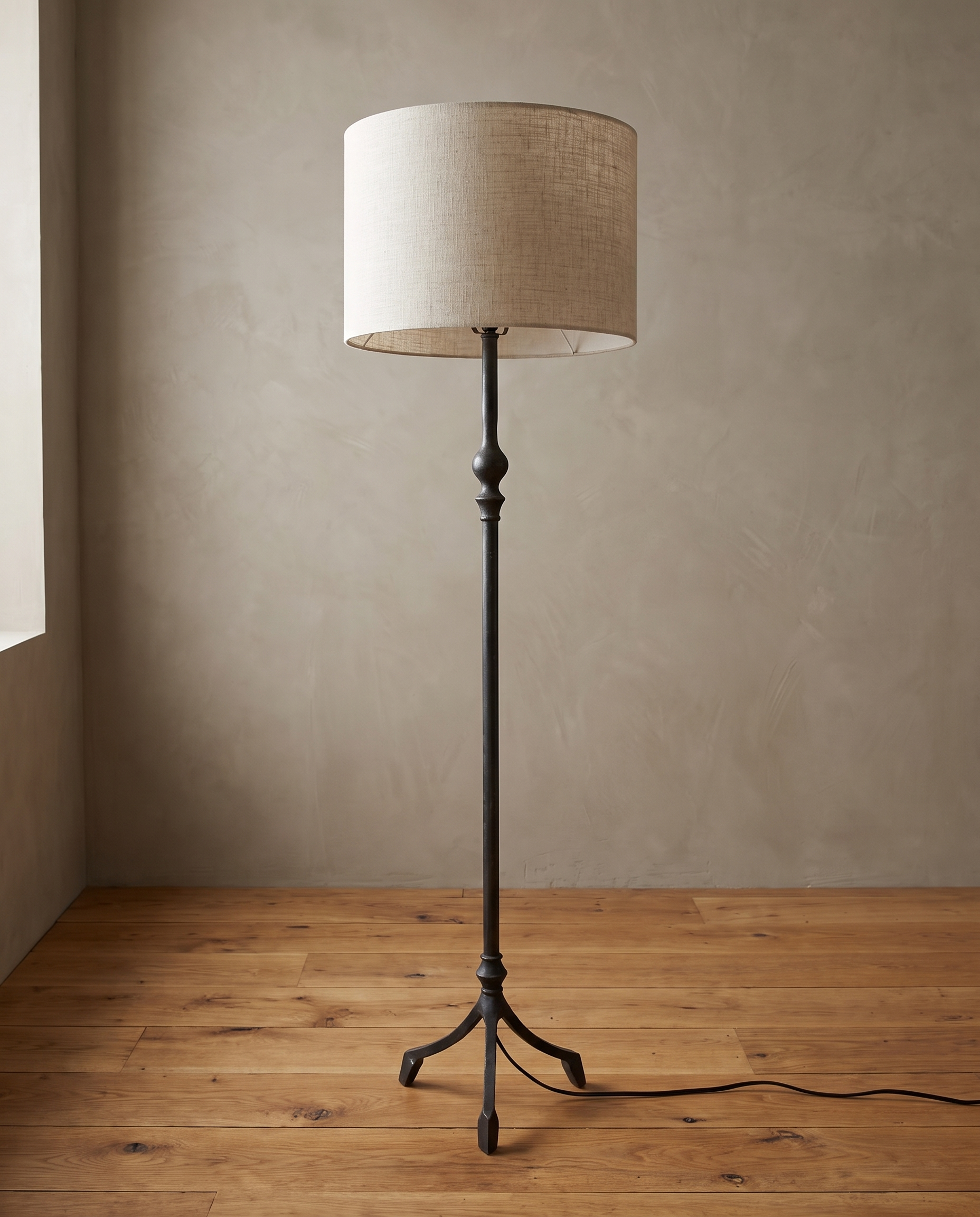 Single forged iron tripod floor lamp at a three-quarter angle, smooth stem with turned knuckle and white linen drum shade