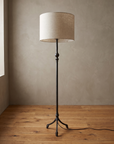 Single forged iron tripod floor lamp at a three-quarter angle, smooth stem with turned knuckle and white linen drum shade