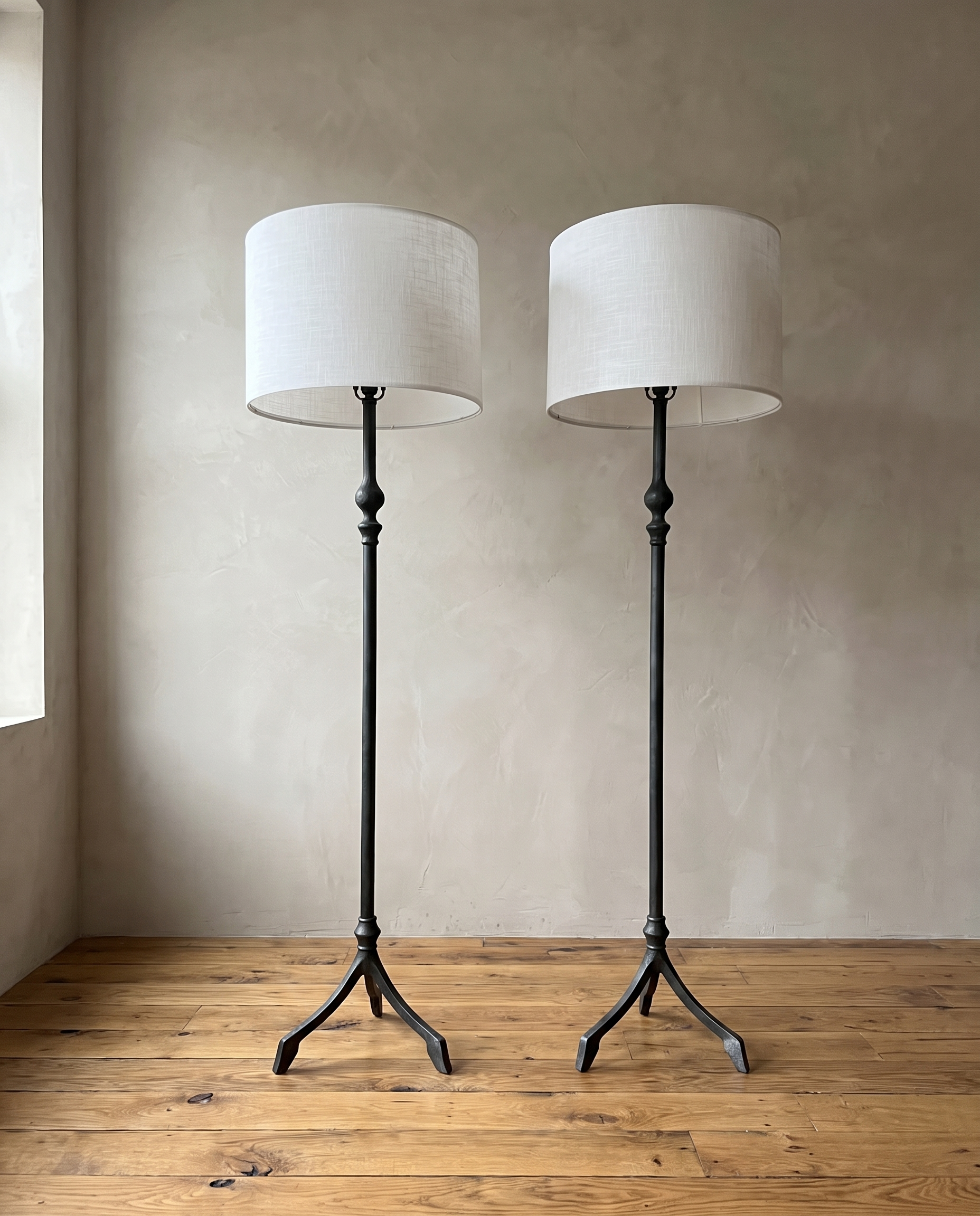 Pair of forged iron tripod floor lamps with white linen drum shades against a greige plaster wall on a reclaimed wood floor