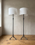 Pair of forged iron tripod floor lamps with white linen drum shades against a greige plaster wall on a reclaimed wood floor