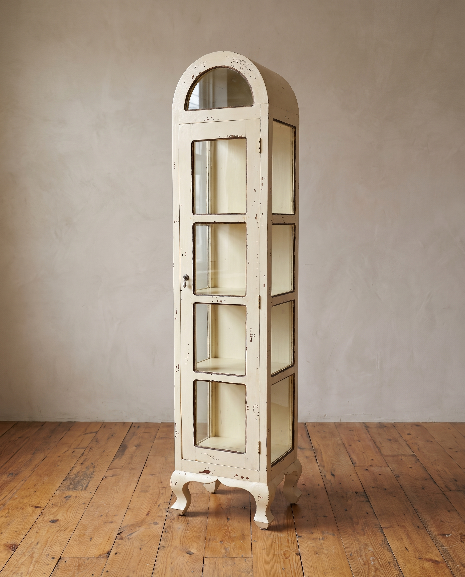 Tall arched glass pantry cabinet in distressed antique cream paint standing against a greige plaster wall on a reclaimed wood floor