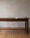 Forged iron tripod floor lamp beside a long antique farmhouse writing desk, plaster wall and reclaimed wood floor