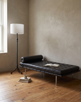 Forged iron tripod floor lamp at the head of a black leather Barcelona daybed in a plaster-walled bedroom