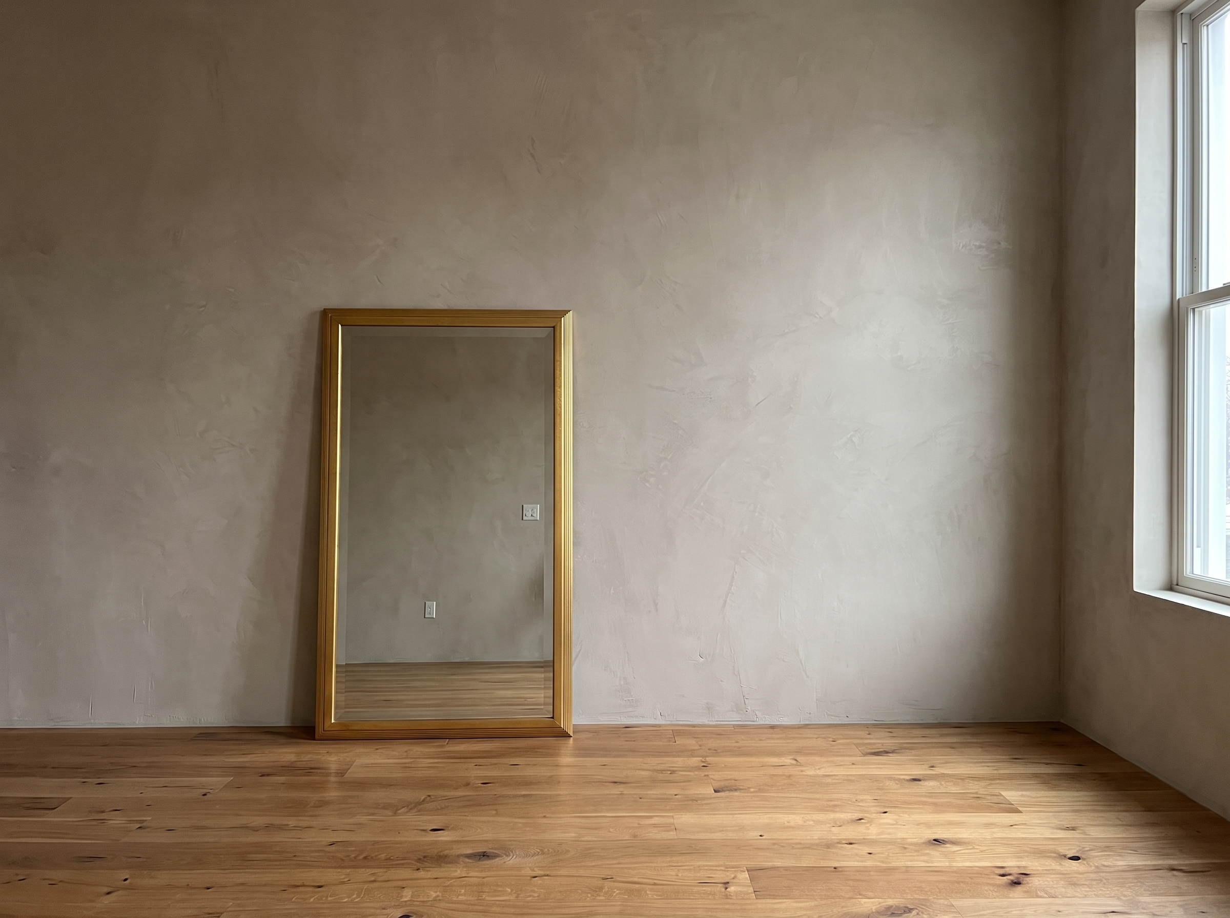 Gilt reeded mirror leaning in a wide plaster room beside a tall window
