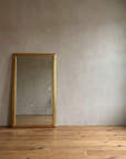 Gilt reeded mirror leaning in a wide plaster room beside a tall window