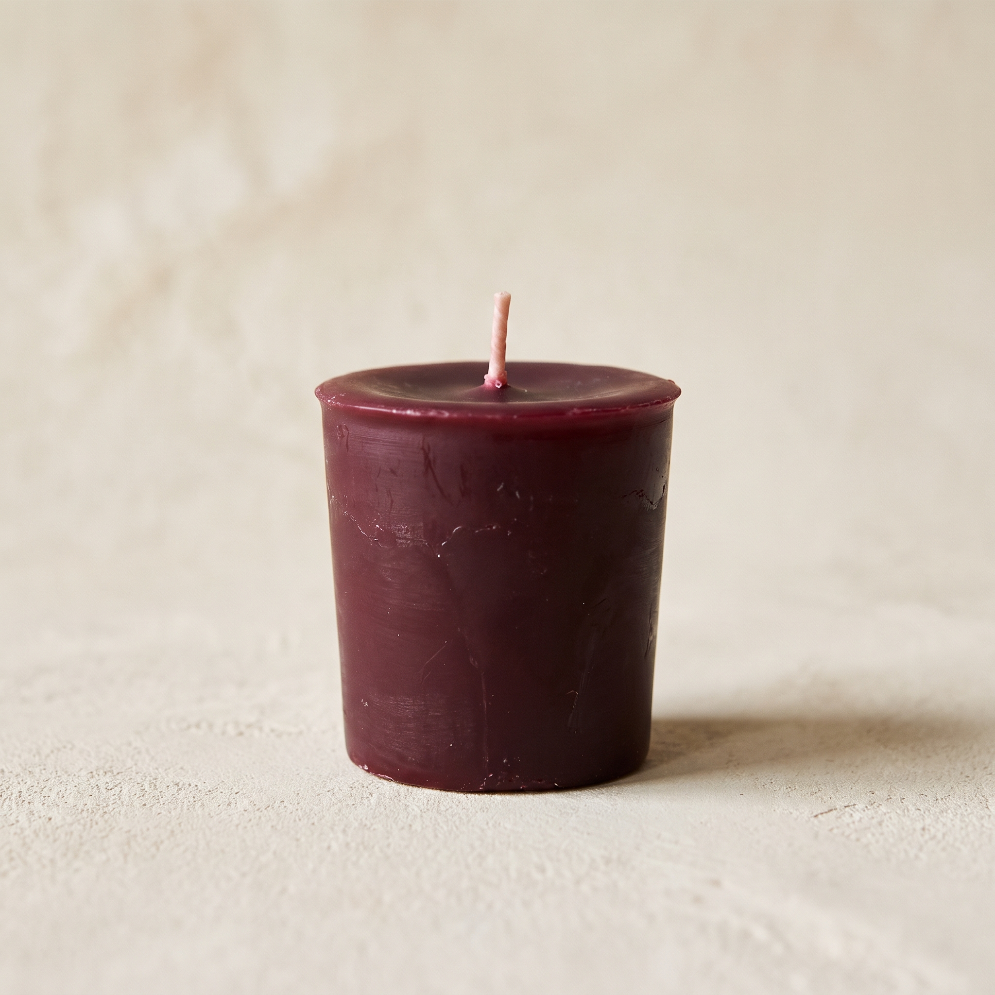 Single burgundy beeswax votive candle on a warm bone plaster background