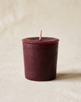 Single burgundy beeswax votive candle on a warm bone plaster background